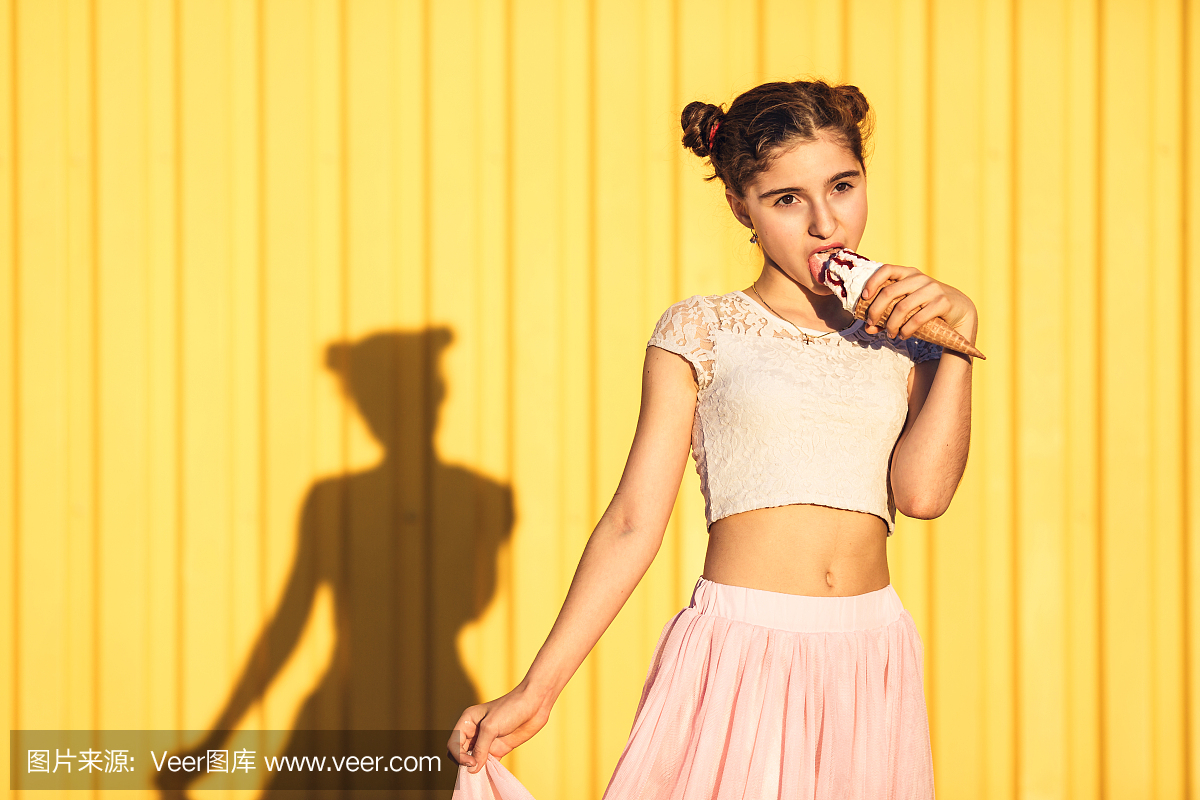 时尚的女孩,在粉红色的裙子舔冰淇淋黄墙。概