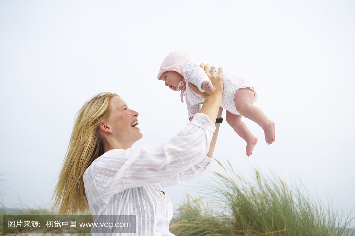 母亲在海滩上举起女婴(0-3个月),微笑