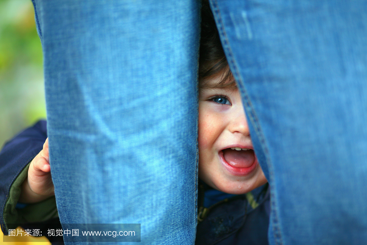 蓝眼睛的男孩盯着母亲的蓝色牛仔裤腿后面