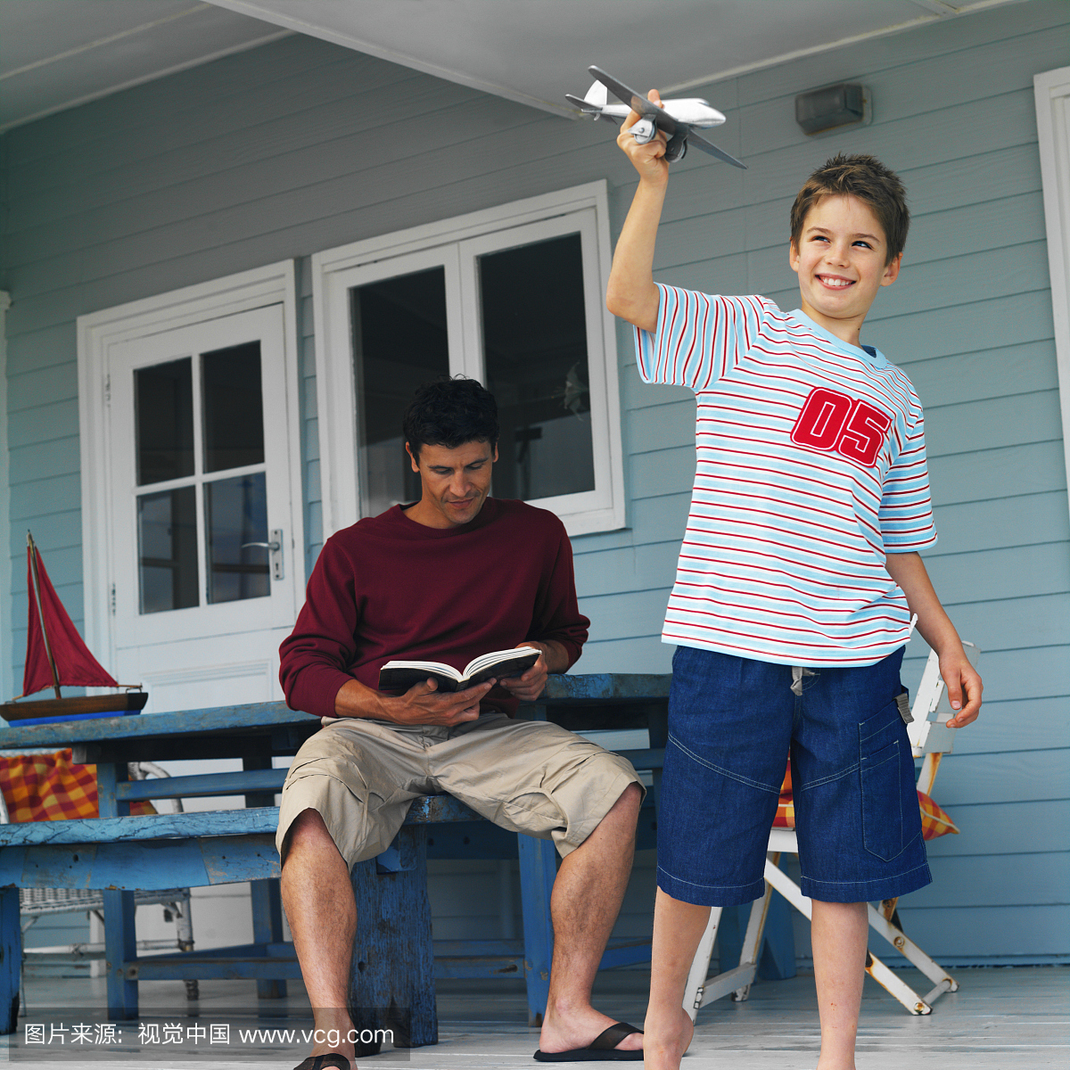 父亲坐阅读书和儿子(8-9)站在飞行玩具飞机